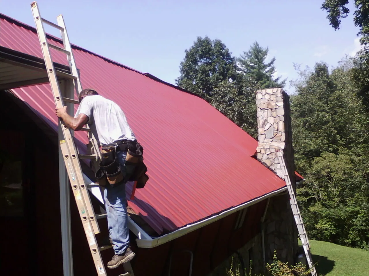 Roofer on a ladder beside a red metal roof and stone chimney — Soffit & Fascia Repair by Roofing Brothers in Wenonah